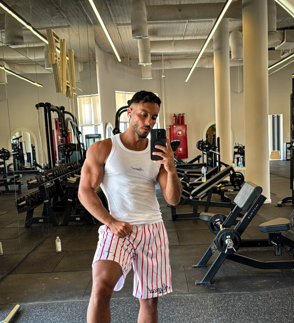 Man taking a mirror selfie in a gym wearing a white tank top and striped shorts.